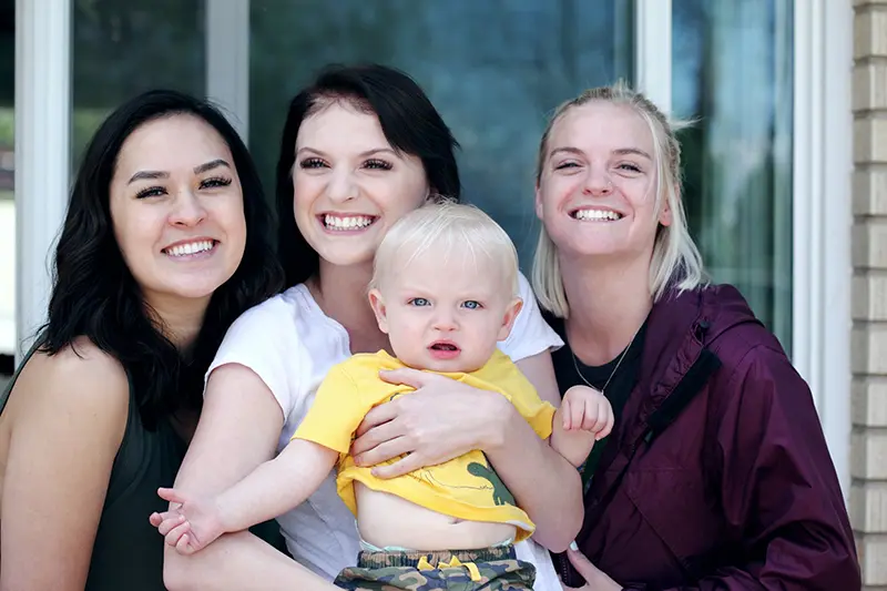 3 women smiling. The middle woman holds a baby